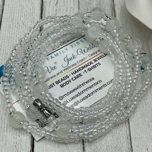 May include: A white beaded waist beads necklace with a silver clasp. The beads are iridescent and have a subtle sparkle. The necklace is wrapped around a white card with text that reads "FAMILY FIRST Vee" Jack-Webb, Business and Wellness Coach | Designe IST BEADS HANDMADE JEWELR BODY CARE T-SHIRTS @mzveewiththetea @Luxeadornmentsco ee@luxeadornments.com".