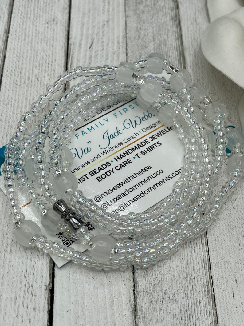 May include: A white beaded waist beads necklace with a silver clasp. The beads are iridescent and have a subtle sparkle. The necklace is wrapped around a white card with text that reads "FAMILY FIRST Vee" Jack-Webb, Business and Wellness Coach | Designe IST BEADS HANDMADE JEWELR BODY CARE T-SHIRTS @mzveewiththetea @Luxeadornmentsco ee@luxeadornments.com".