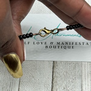 May include: A black beaded necklace with a gold clasp. The necklace is on a white surface with the text "Self Love & Manifestation Boutique".