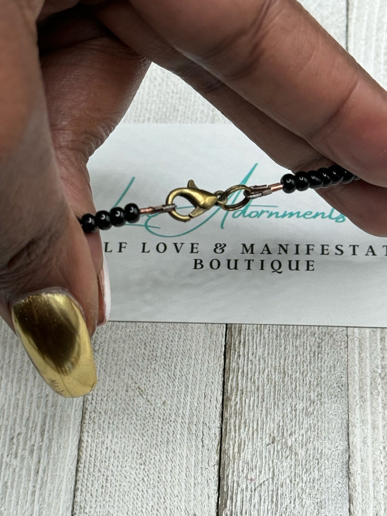 May include: A black beaded necklace with a gold clasp. The necklace is on a white surface with the text "Self Love & Manifestation Boutique".