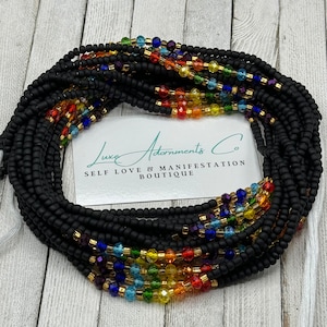 May include: A black waist bead necklace with rainbow coloured glass beads and gold accents. The beads are arranged in a rainbow pattern. The necklace is on a wooden surface. "Luxe Adornments Co. Self Love & Manifestation Boutique" is printed on a white card.