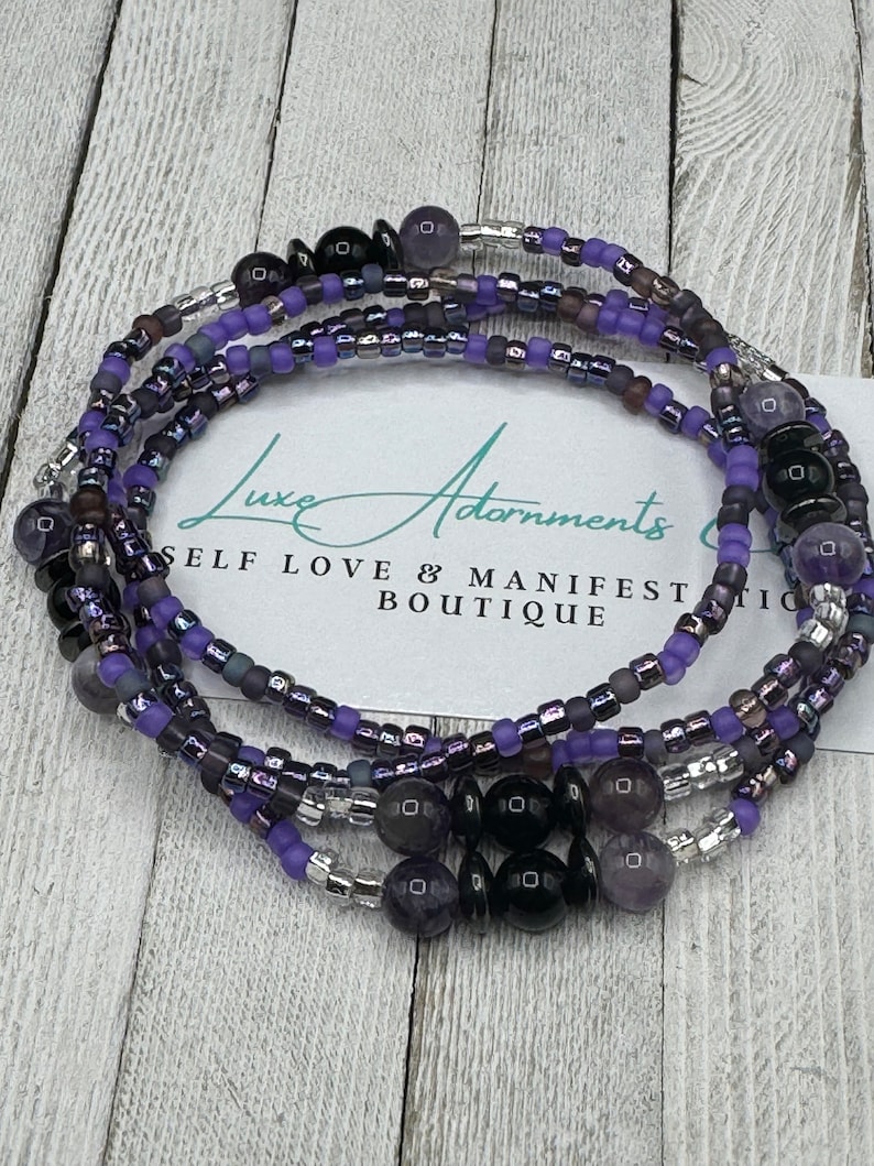 May include: Purple and black beaded waist beads with a silver accent. The beads are arranged in a layered pattern and include small round beads and larger round beads. The text "Luxe Adornments Self Love & Manifest Boutique" is visible in the background.