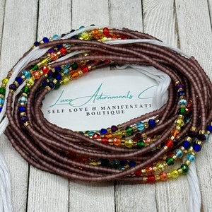 May include: A collection of beaded waist beads in shades of purple, with strands of colorful beads in rainbow hues. The beads are strung on white string. The text "Luxe Adornments Co. Self Love & Manifestation Boutique" is visible.
