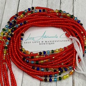 May include: A red beaded waist beads with colorful beads woven throughout. The beads are arranged in a circle and are on a wooden surface. The text "Luxe Adornments Self Love & Manifestation Boutique" is visible on a white card.