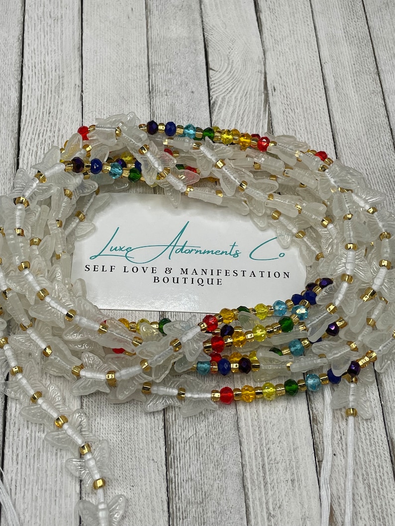 Puede incluir: Cuentas de cintura blancas con dijes de mariposa y cuentas de acento coloridas. Las cuentas de la cintura est&aacute;n ensartadas en un cord&oacute;n blanco y presentan detalles dorados. La imagen incluye una tarjeta con el texto "Luxe Adornments Co. SELF LOVE & MANIFESTATION BOUTIQUE."
