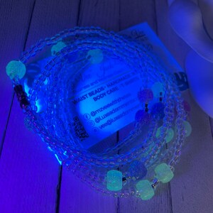 May include: A set of clear waist beads with glow-in-the-dark beads. The beads are arranged in a circle and are strung on a clear elastic cord. The beads are a variety of shapes and sizes, and some of them have a sparkly finish. The beads are a light blue color in the dark.