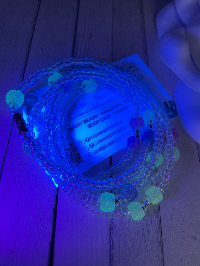 May include: A set of clear waist beads with glow-in-the-dark beads. The beads are arranged in a circle and are strung on a clear elastic cord. The beads are a variety of shapes and sizes, and some of them have a sparkly finish. The beads are a light blue color in the dark.