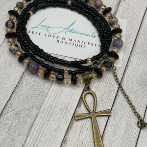 May include: A black beaded waist chain with gold and purple beads, an ankh pendant, and the text "Luxe Adornments Self Love & Manifest Boutique".