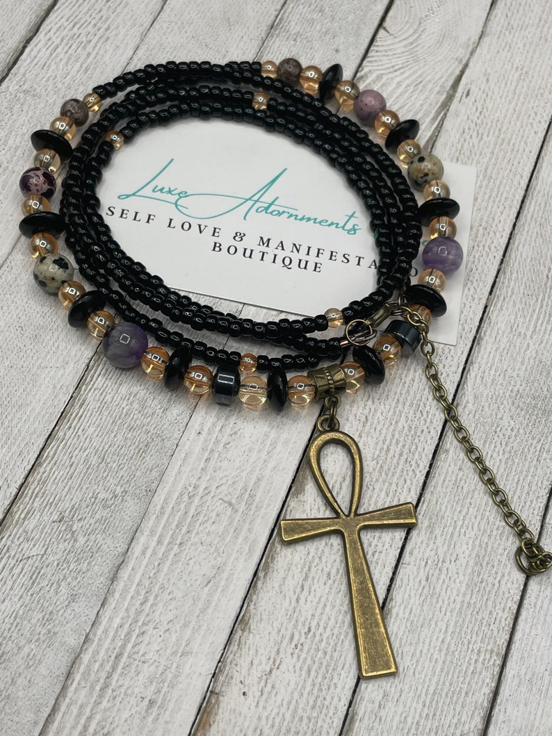 May include: A black beaded waist chain with gold and purple beads, an ankh pendant, and the text "Luxe Adornments Self Love & Manifest Boutique".