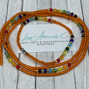 K&ouml;nnte beinhalten: Eine mehrstr&auml;ngige Taillenperlenkette mit orangefarbenen, goldenen, blauen, gr&uuml;nen und roten Perlen. Die Perlen sind in einem Regenbogenmuster angeordnet. Die Kette liegt auf einer Holzoberfl&auml;che mit einer Visitenkarte, auf der "Luxe Adornments Co. Self Love & Manifestation Boutique" steht.