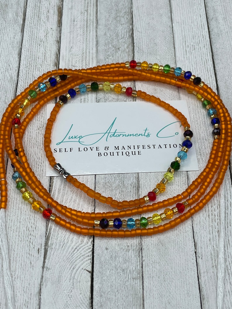 K&ouml;nnte beinhalten: Eine mehrstr&auml;ngige Taillenperlenkette mit orangefarbenen, goldenen, blauen, gr&uuml;nen und roten Perlen. Die Perlen sind in einem Regenbogenmuster angeordnet. Die Kette liegt auf einer Holzoberfl&auml;che mit einer Visitenkarte, auf der "Luxe Adornments Co. Self Love & Manifestation Boutique" steht.