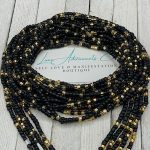 May include: Black and gold waist beads. The beads are a mix of black and gold, strung together to create a layered effect. The text "Luxe Adornments Co. SELF LOVE & MANIFESTATION BOUTIQUE" is visible on a white card.