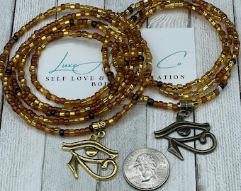 May include: Two gold-toned Egyptian eye of Horus charms hanging from a beaded waist chain. The chain is made of brown and gold beads and is layered. The charms are hanging from the chain with a small loop. The charms are in focus and the chain is slightly out of focus. The background is a light wood surface.