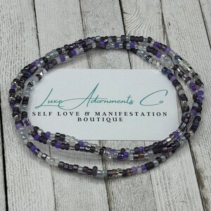 May include: A multi-strand beaded necklace with purple, gray, and silver beads. The necklace is on a wooden surface with a business card that reads "Luxe Adornments Co. Self Love & Manifestation Boutique".