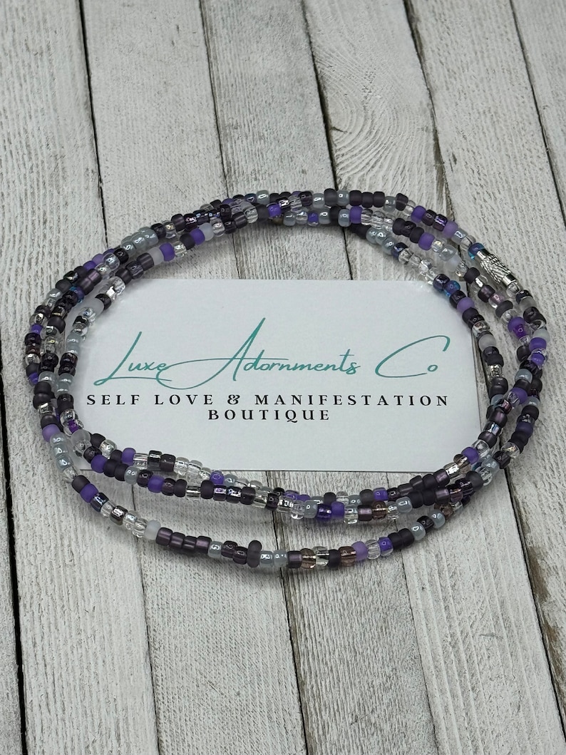 May include: A multi-strand beaded necklace with purple, gray, and silver beads. The necklace is on a wooden surface with a business card that reads "Luxe Adornments Co. Self Love & Manifestation Boutique".