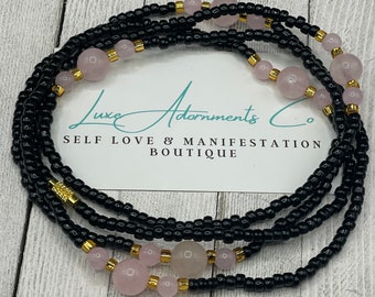 May include: A black beaded waist bead with rose quartz beads and gold accents. The beads are arranged in a circular pattern and are strung on a black cord. The text "Luxe Adornments Co. Self Love & Manifestation Boutique" is visible on a white card behind the beads.