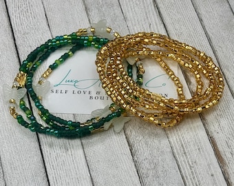 May include: Two strands of waist beads. One is gold, the other is green with gold accents and white butterfly charms. A white card with the text "Luxe Self Love & Bout" is in the background.