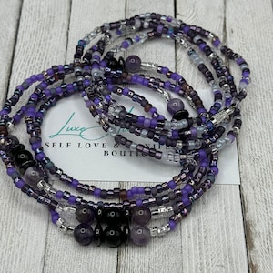 May include: A multi-strand beaded bracelet with purple, silver, and black beads. The bracelet is made of several strands of beads, including small glass beads, larger amethyst beads, and black onyx beads. The bracelet is tied with a knot and has a "Self Love & Life Boutique" tag attached.