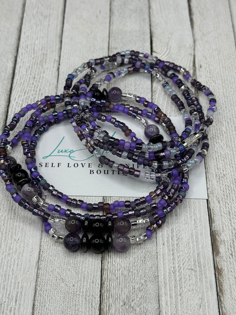 May include: A multi-strand beaded bracelet with purple, silver, and black beads. The bracelet is made of several strands of beads, including small glass beads, larger amethyst beads, and black onyx beads. The bracelet is tied with a knot and has a "Self Love & Life Boutique" tag attached.