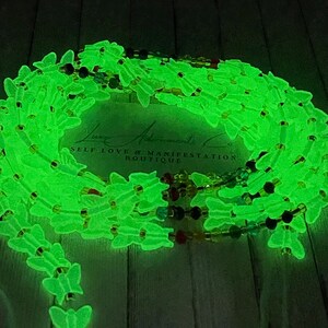 Puede incluir: Cuentas de cintura que brillan en la oscuridad con dijes de mariposa. Las cuentas son de color verde ne&oacute;n brillante y est&aacute;n ensartadas con cuentas m&aacute;s peque&ntilde;as y coloridas. El texto "Luxe Adornments Co. Self Love & Manifestation Boutique" es visible.