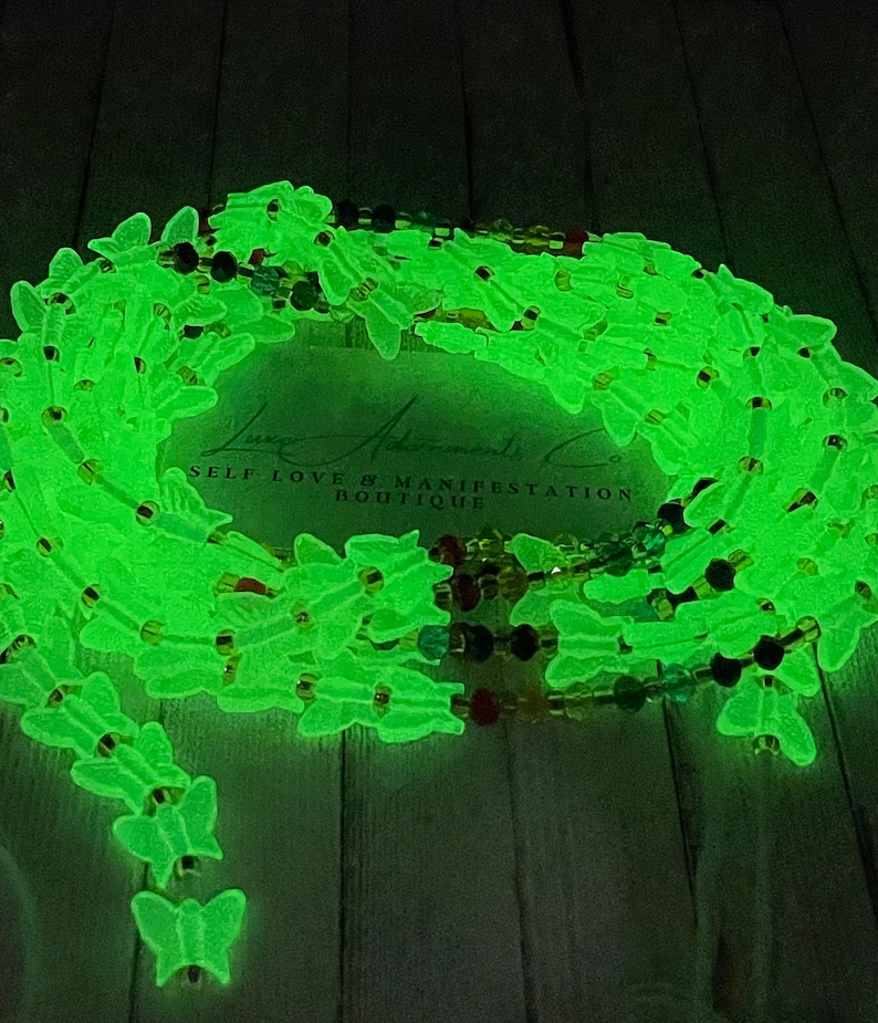 Puede incluir: Cuentas de cintura que brillan en la oscuridad con dijes de mariposa. Las cuentas son de color verde ne&oacute;n brillante y est&aacute;n ensartadas con cuentas m&aacute;s peque&ntilde;as y coloridas. El texto "Luxe Adornments Co. Self Love & Manifestation Boutique" es visible.