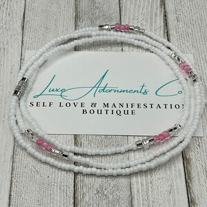 May include: White waist beads with pink beads and silver accents. The beads are arranged in three rows and are tied together with a silver clasp. The beads are on a white background with the text "Luxe Adornments Co. Self Love & Manifestation Boutique".