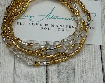 May include: A gold and clear beaded waist bead with a silver clasp. The beads are arranged in a layered pattern. The waist bead is on a wooden surface. "Luxe Adornments Self Love & Manifest Boutique" is written on a white card behind the waist bead.