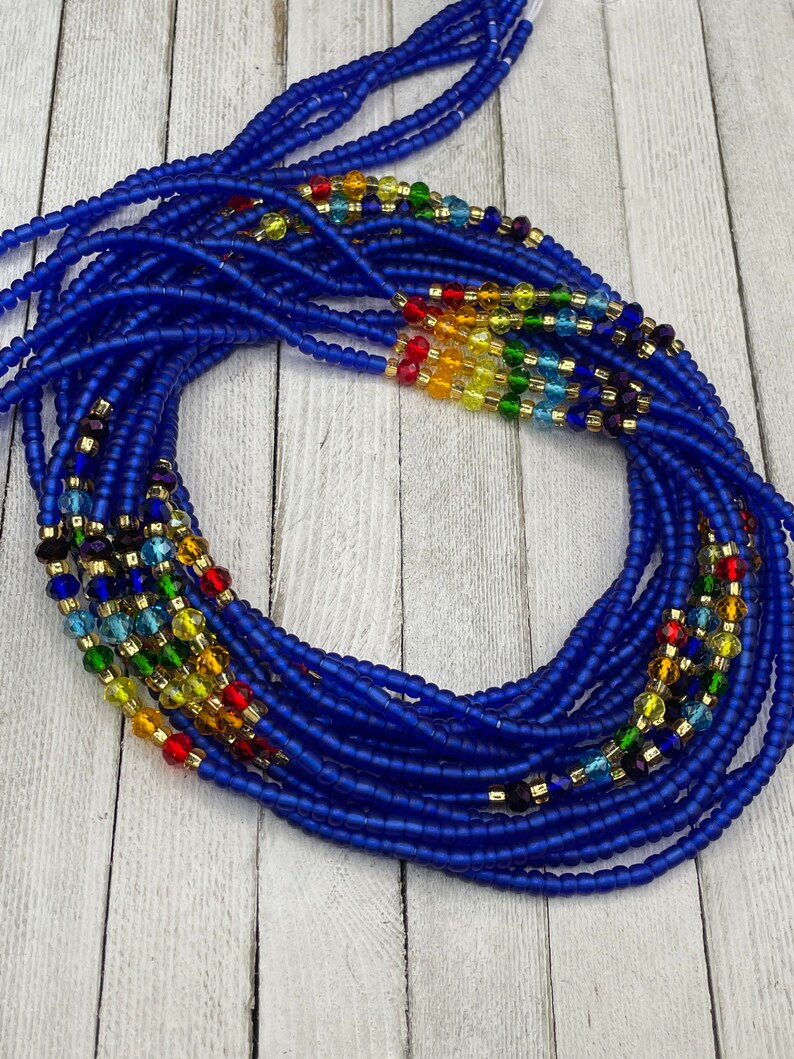 May include: A collection of blue waist beads with colorful accents. The beads are a deep blue, with sections of rainbow-colored beads and gold-colored spacers. The beads are arranged in a circular pattern, suggesting they are ready to wear.