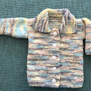 May include: A long-sleeved cardigan made of fuzzy, variegated yarn in shades of light blue, yellow, cream, and grey. The garment features a wide collar and a front closure secured by five round, white, textured buttons arranged in a vertical line.