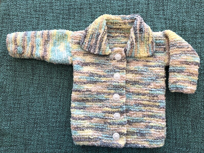 May include: A long-sleeved cardigan made of fuzzy, variegated yarn in shades of light blue, yellow, cream, and grey. The garment features a wide collar and a front closure secured by five round, white, textured buttons arranged in a vertical line.