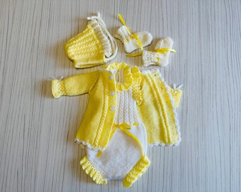 May include: A set of knitted baby clothes in yellow and white, including a romper, cardigan, hat, and booties. The romper and cardigan have yellow trim and button closures. The booties and hat are accented with yellow ribbons.