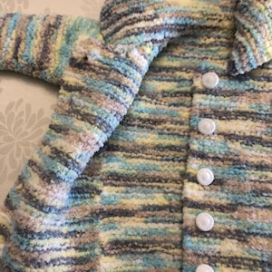 May include: A long-sleeved cardigan made of fuzzy, variegated yarn in shades of light blue, pale yellow, white, and grey. The garment features a fold-over collar and a vertical row of five round, white textured buttons along the front opening.
