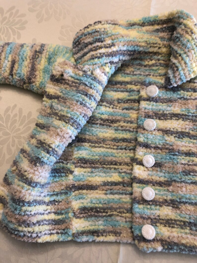 May include: A long-sleeved cardigan made of fuzzy, variegated yarn in shades of light blue, pale yellow, white, and grey. The garment features a fold-over collar and a vertical row of five round, white textured buttons along the front opening.