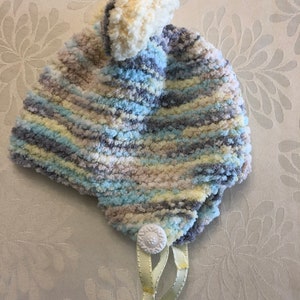 May include: A soft, fuzzy knit hat featuring a variegated color pattern of light blue, cream, grey, and pale yellow. The hat is topped with a large, round pom-pom and includes a white decorative button at the base with two light yellow ribbons.