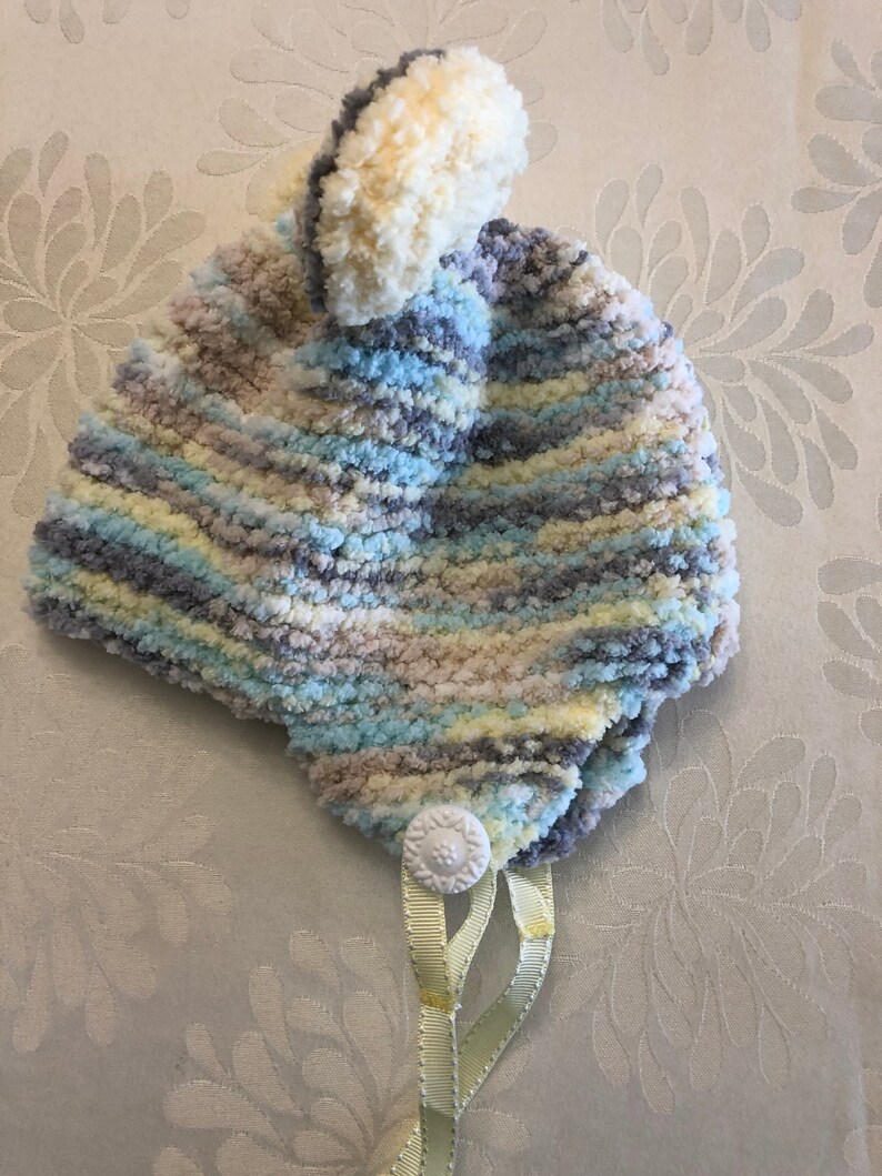 May include: A soft, fuzzy knit hat featuring a variegated color pattern of light blue, cream, grey, and pale yellow. The hat is topped with a large, round pom-pom and includes a white decorative button at the base with two light yellow ribbons.