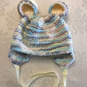 May include: A soft, fuzzy, variegated yarn hat featuring two rounded bear-like ears on top. The hat is knitted in a blend of light blue, grey, white, and cream colors. It includes two light yellow ribbon ties attached to the bottom ear flaps.