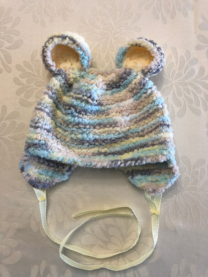May include: A soft, fuzzy, variegated yarn hat featuring two rounded bear-like ears on top. The hat is knitted in a blend of light blue, grey, white, and cream colors. It includes two light yellow ribbon ties attached to the bottom ear flaps.