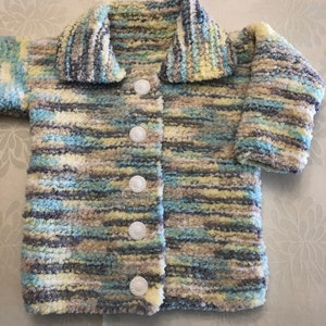 May include: A long-sleeved cardigan made of fuzzy, variegated yarn in shades of light blue, pale yellow, grey, and white. The garment features a wide collar, long sleeves, and a front closure secured by five round, white, textured buttons.