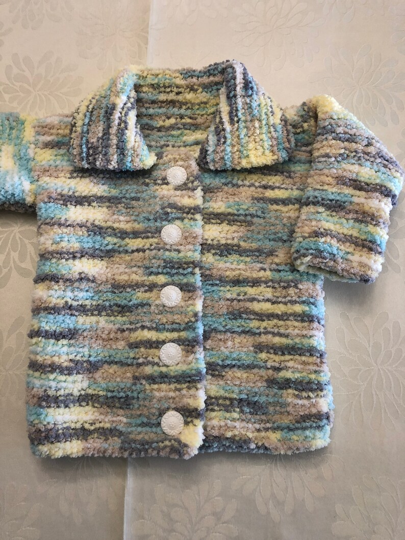 May include: A long-sleeved cardigan made of fuzzy, variegated yarn in shades of light blue, pale yellow, grey, and white. The garment features a wide collar, long sleeves, and a front closure secured by five round, white, textured buttons.