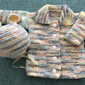 May include: A long-sleeved cardigan and a matching hat with bear ears, both crafted from a soft, variegated yarn in shades of yellow, light blue, grey, and white. The cardigan features a wide collar and four white buttons down the front.