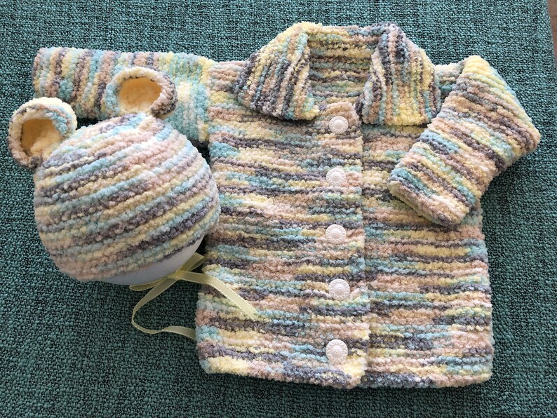 May include: A long-sleeved cardigan and a matching hat with bear ears, both crafted from a soft, variegated yarn in shades of yellow, light blue, grey, and white. The cardigan features a wide collar and four white buttons down the front.