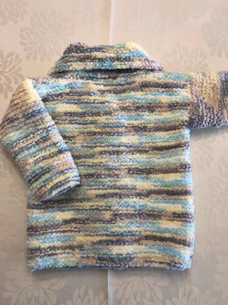 May include: A long-sleeved sweater crafted from fuzzy, textured yarn featuring a variegated color palette of soft yellow, light blue, white, and grey. The garment includes a wide, folded collar and a straight hemline, showcasing a consistent horizontal striped pattern throughout the entire piece.