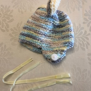 May include: A soft, fuzzy knit hat featuring a variegated color pattern of light blue, cream, gray, and white. The hat has a small pom-pom on top, ear flaps, a single white button on the side, and two long, pale yellow ribbon ties attached.