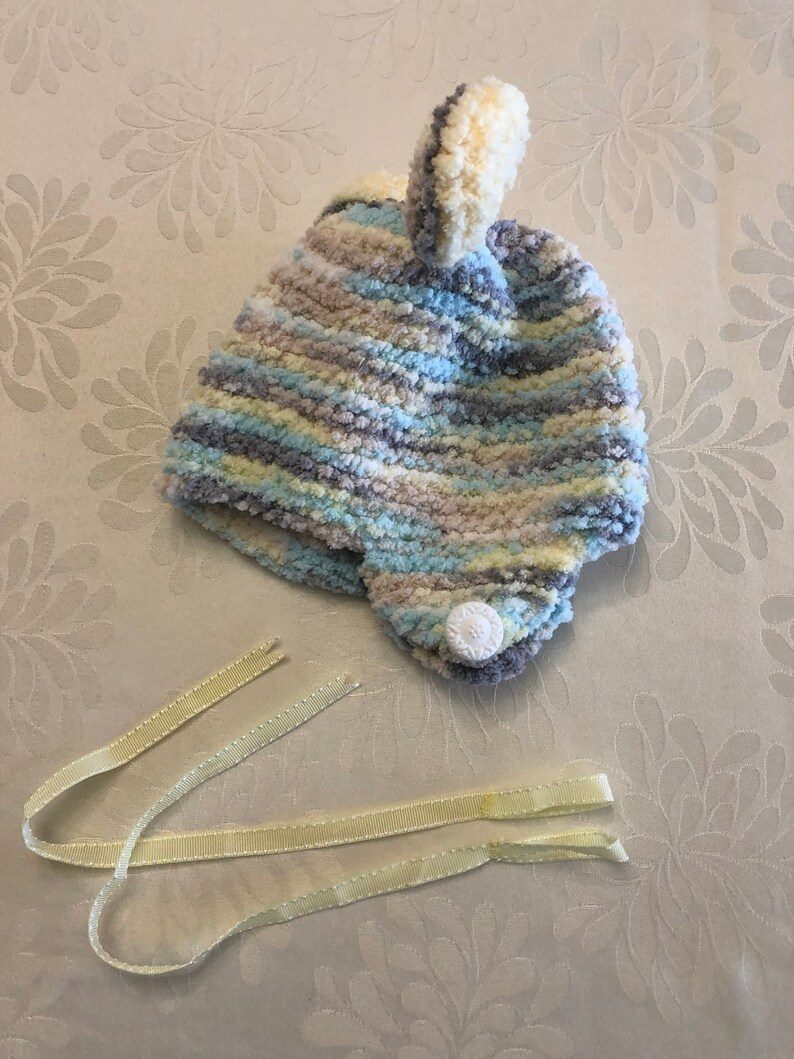 May include: A soft, fuzzy knit hat featuring a variegated color pattern of light blue, cream, gray, and white. The hat has a small pom-pom on top, ear flaps, a single white button on the side, and two long, pale yellow ribbon ties attached.