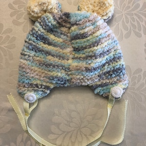 May include: A knit hat featuring a variegated yarn pattern in shades of blue, gray, cream, and yellow. The hat includes two round, textured pom-poms on top, two white decorative buttons at the base, and two light yellow ribbons for fastening.