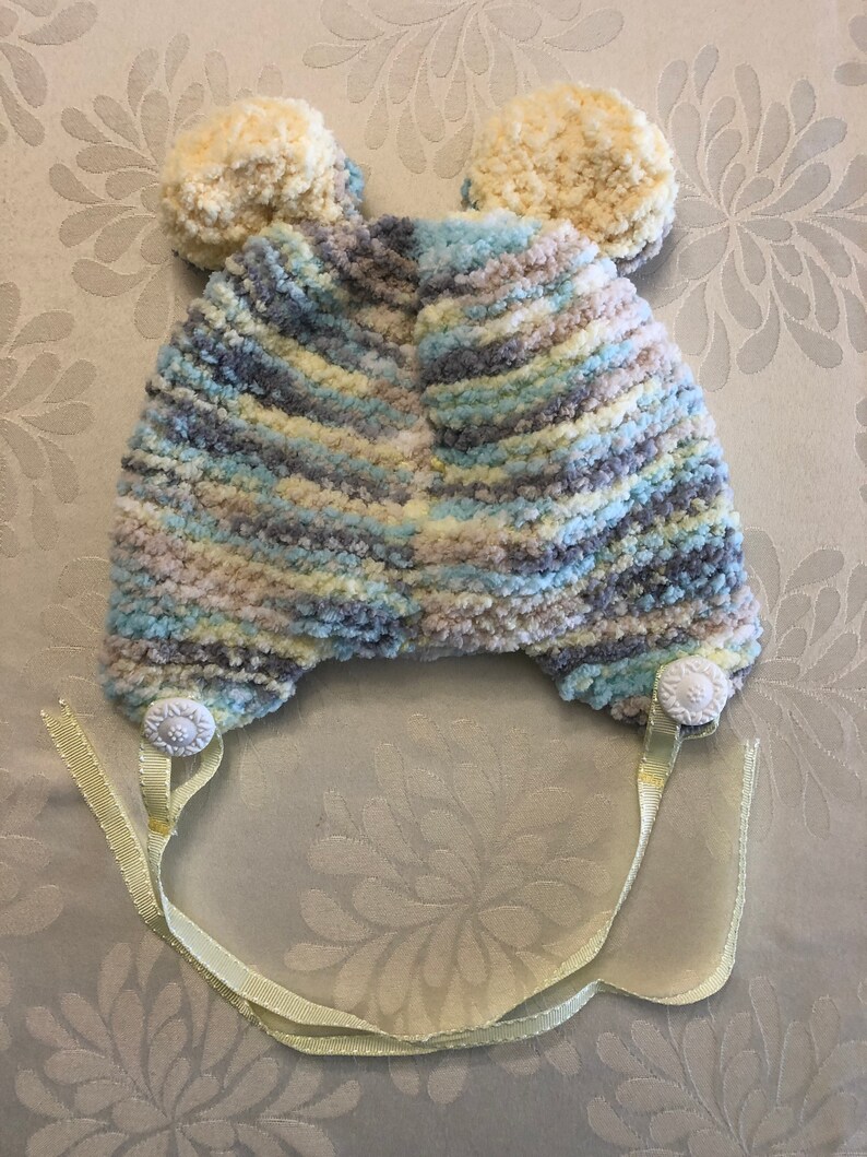May include: A knit hat featuring a variegated yarn pattern in shades of blue, gray, cream, and yellow. The hat includes two round, textured pom-poms on top, two white decorative buttons at the base, and two light yellow ribbons for fastening.