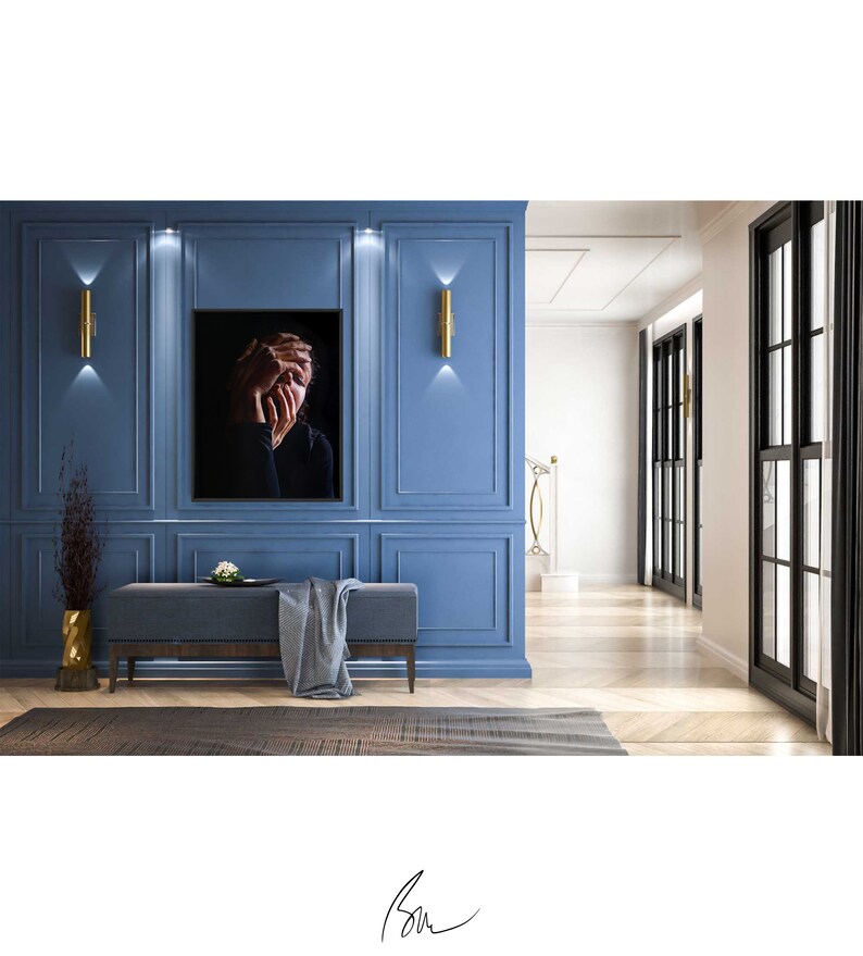 May include: A black and white photograph of a person with their hands covering their face, hanging on a blue wall in a modern interior design setting. The photograph is framed and hangs above a gray bench with a gray throw blanket.