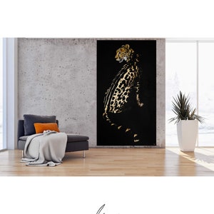 Op de afbeelding: Een zwart en gouden luipaardprint schilderij op een zwarte achtergrond. De luipaard zit en kijkt naar links. Het schilderij is ingelijst en hangt aan een muur in een kamer met een bank en een plant.