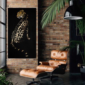 Op de afbeelding: Een zwart en gouden canvas wandkunstprint met luipaardprint, hangend aan een bakstenen muur boven een bruine leren fauteuil en voetenbank.