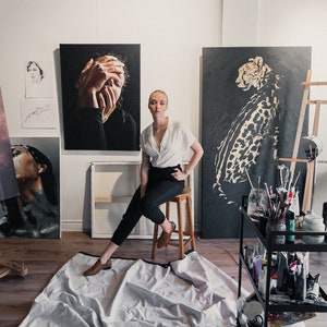 May include: A woman wearing a white shirt and black pants sits on a stool in an art studio. She is surrounded by paintings, including a portrait of a woman with her hands covering her face, a cheetah, and a nude figure. The studio is well-lit and has a wooden floor.